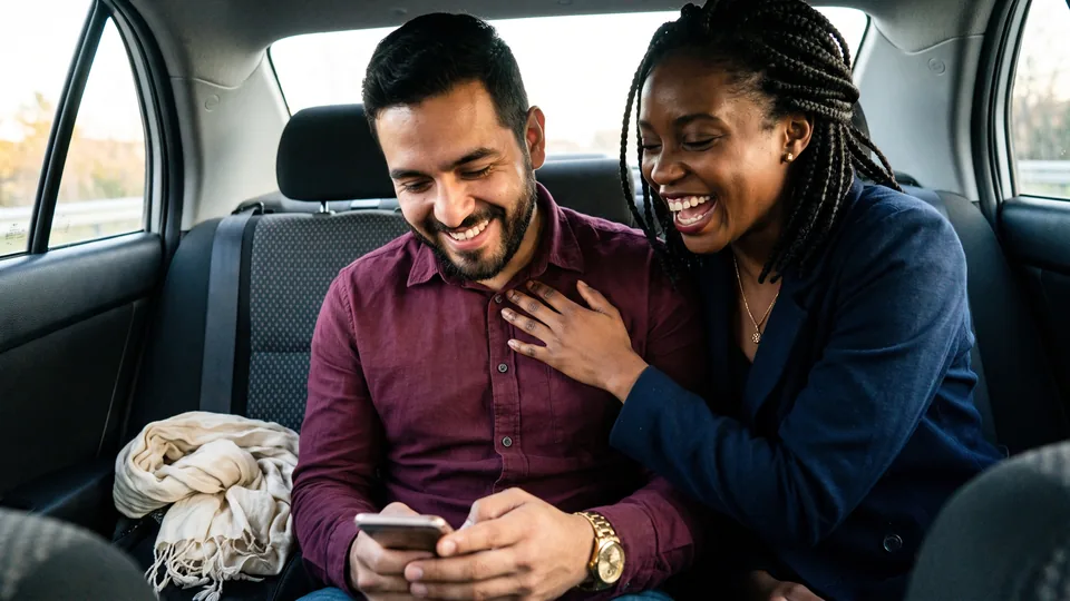 A couple in the back seat of a car, laughing together at something on his phone